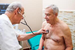 An elderly man sits on the examination table, his hands resting on his lap as the doctor listens to his heartbeat with a stethoscope. photo