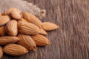 A close-up of raw almonds scattered on a wooden surface, showcasing their natural texture and color, with a burlap sack partially visible in the background. photo