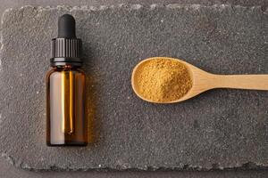 A dropper bottle and a spoonful of powder sit on a textured stone surface, suggesting a focus on natural remedies or essential oils. photo