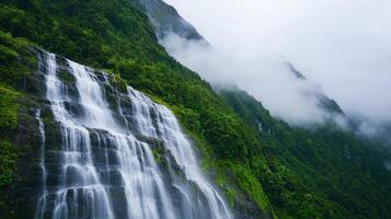 A stunning waterfall cascades down a lush, green mountain, surrounded by misty clouds and vibrant vegetation. photo