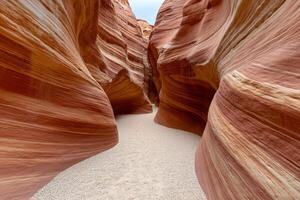 A narrow, winding canyon features stunning red and orange rock formations, with smooth, layered textures and a sandy pathway through the dramatic landscape. photo