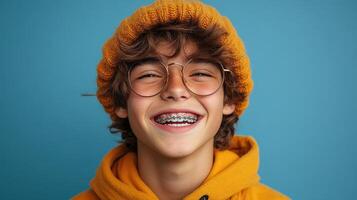 Young boy in bright orange beanie and glasses smiles against a blue background, showcasing a joyful expression photo