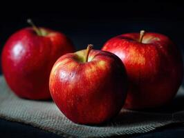 Three shiny red apples with dew droplets rest on a piece of burlap, showing varied hues from deep red to yellowish-orange. The surface texture is smooth and fresh photo