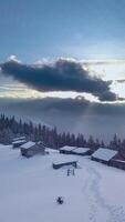 Snow blankets rustic cabins in a serene winter landscape. Dark clouds loom above, contrasting with soft sunlight breaking through, creating a tranquil atmosphere. Vertical footage video