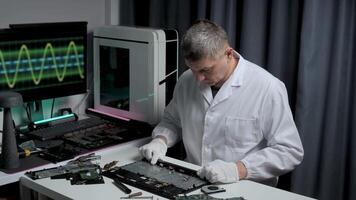 A technician in a white coat focuses on repairing a circuit board on a workbench. Monitors display data while tools are scattered around in a high-tech lab. video