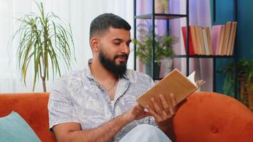 Indian man reading interesting book turning pages smiling enjoying literature taking a rest on sofa video