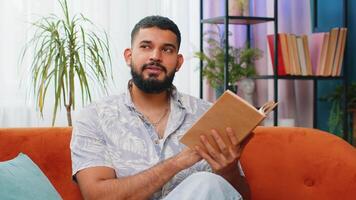 Indian man reading interesting book turning pages smiling enjoying literature taking a rest on sofa video