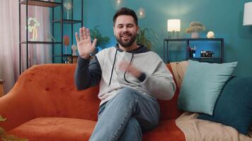 Caucasian man smiling friendly at camera, waving hands gesturing hello, hi, greeting at home on sofa video