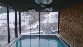 Modern indoor pool with wooden walls and soft lighting, overlooking snowy mountains, a ski slope, and trees through large windows in the Dolomites. video
