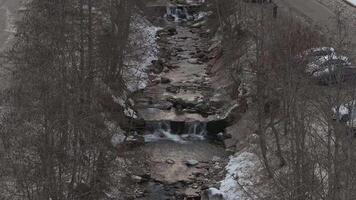 Aerial view of a mountain stream flowing through a rocky channel with a small waterfall, leafless trees, a road, and light snow in the Dolomites. video