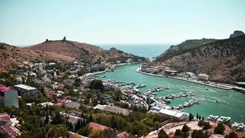 Panoramic view of coastal town nestled in valley, with marina and sea in background. Shot on sunny day from high angle. Camera pans right, revealing town's layout and surrounding landscape. video