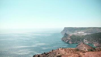 Sweeping panoramic view of a rugged coastline meeting the vast ocean, shot from a high vantage point on a sunny day. Camera pans left, revealing the dramatic cliffs and the serene sea. video