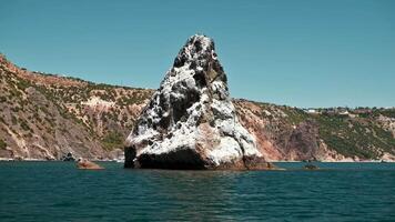 Rock formation rises dramatically from turquoise sea waters, surrounded by a coastal scene of rugged cliffs and sparse vegetation. daylight with a clear sky. video