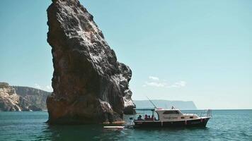 un pequeño barco anclado junto a un imponente rocoso acantilado en un tranquilo mar. claro cielo, mientras varios personas son visible en bote. natural belleza de escabroso acantilados, evoca sentido de aventuras y relajación. video