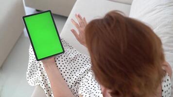 Red-Haired Woman Sitting on Couch in Polka Dot Dress Using Tablet with Green Screen, Touching Display, Relaxing in Living Room, Digital Interaction, Modern Technology, Smart Device and Home Leisure. video