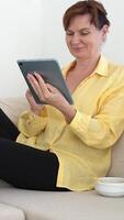 Middle-Aged Woman Sitting on Couch Using Tablet, Thoughtfully Reading Digital Content and Eats Junk Food in Casual Home Setting, Relaxed Atmosphere, Comfortable Outfit, Leisure and Technology Concept. video
