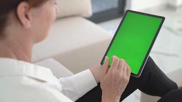 Middle-Aged Woman Using Tablet With Green Screen, Sitting on Sofa in Bright Living Room, Tapping and Scrolling on Touchscreen, Digital Device Interaction, Wireless Technology and Home Comfort. video