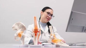 An orthopedic specialist works in a modern office using a computer. Models of the knee joint, pelvic floor and foot are on the table. Concept of orthopedic diagnostics video