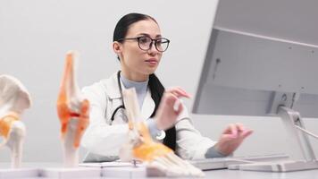 An orthopedic specialist works in a modern office using a computer. Models of the knee joint, pelvic floor and foot are on the table. Concept of orthopedic diagnostics. video