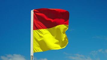 A red and yellow flag waves in the wind against a blue sky, marking designated swimming zones on Australian beaches. Essential coastal safety symbol protecting beachgoers in surf conditions. video