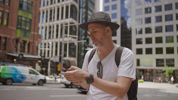 A man in a black hat, white t-shirt, and backpack is standing in an urban environment, focused on his smartphone. He appears to be using navigation video