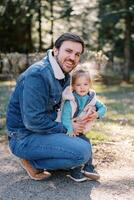 Smiling dad squatting and hugging little daughter in park photo