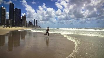 A middle-aged man runs joyfully along the shoreline at Gold Coast beach, kicking up ocean spray as towering skyscrapers frame the cloud-dotted sky. video