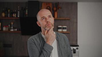 A middle-aged man in casual clothes, standing in his kitchen. He looks deep in thought, contemplating different options and struggling to make a decision. video