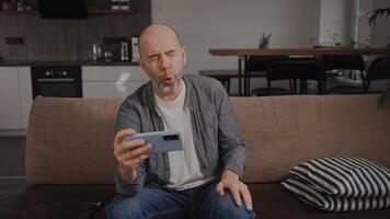 uma meia idade homem às casa em a sofá, rindo Fora alto enquanto assistindo muito engraçado videos em dele Smartphone. ele s desfrutando entretenimento conteúdo a partir de social meios de comunicação, imerso dentro a Diversão do a Internet.