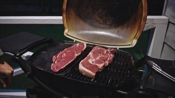 Two raw steaks are sizzling on a gas grill, illuminated by warm light at night. The barbecue lid is open, revealing the marbled texture of the meat as it starts to cook over the open flame. video