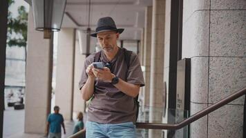 A middle-aged man wearing a hat and backpack stands in the city, using his smartphone to check directions, news, and mobile apps. Urban traveler navigating with digital technology on a bright day. video