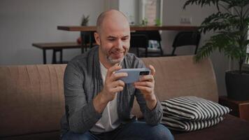 An expressive middle-aged man is passionately playing a game on his smartphone. His face shows excitement and deep engagement. He is at home, sitting on a cozy sofa video