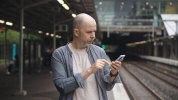 A middle-aged man stands on a city train platform checking the electric train schedule on his smartphone. This urban commuter moment showcases modern digital convenience within transport systems. video