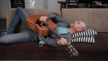 A man lies back on a soft couch with an acoustic guitar resting on his chest. He appears relaxed, possibly dozing off or taking a break from playing. video