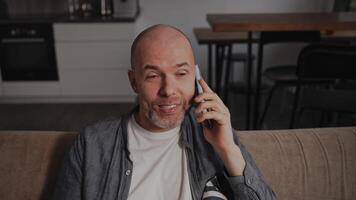 An adult man sits at home on the sofa, talking on the phone and laughing. The middle-aged man is showing bright, positive emotions and is in a good mood. At home video