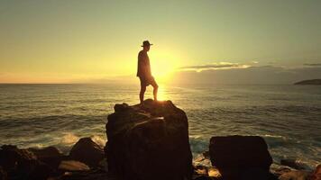 The silhouette of a man in a hat stands atop a large oceanfront rock against a breathtaking sunrise sunset sky. This inspiring seascape captures the magical transition between night and day video