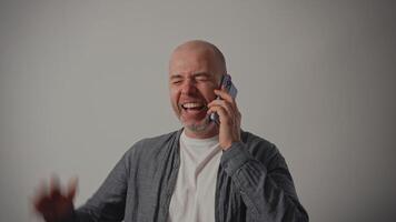 A middle-aged man in casual clothes stands against a white wall, laughing during a phone conversation. He s in a good mood, enjoying the conversation with a friend, showing a great sense of humor. video