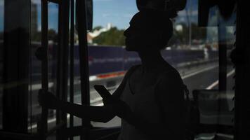 Silhouette of young woman standing inside moving city bus, holding handrail while using smartphone during commute. Through windows, visible traffic on busy highway. video