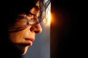 Woman gazing thoughtfully at sunset through a window with soft light illuminating her face photo