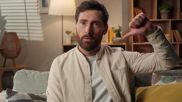 Sad frowning upset man Caucasian dissatisfied guy male home owner on couch showing thumb down gesture finger dislike hand gesture bad sign disapprove no negative result displeased looking at camera video