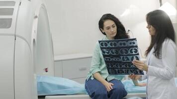Woman doctor radiologist explains the results of CT scanning for young female patient, showing the snapshot with images, observing and analyzing CT scan in modern clinic beside modern CT scanner 24 video