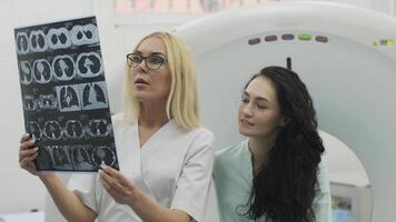 Woman doctor radiologist explains the results of CT scanning for young female patient, showing the snapshot with images, observing and analyzing CT scan in modern clinic beside modern CT scanner 4 video