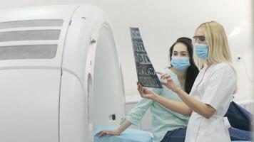 Woman doctor radiologist explains the results of CT scanning for young female patient, showing the snapshot with images, observing and analyzing CT scan in modern clinic beside modern CT scanner 16 video