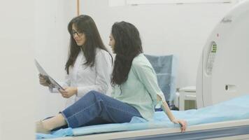 Woman doctor radiologist explains the results of CT scanning for young female patient, showing the snapshot with images, observing and analyzing CT scan in modern clinic beside modern CT scanner 20 video