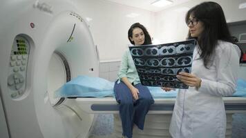 Woman doctor radiologist explains the results of CT scanning for young female patient, showing the snapshot with images, observing and analyzing CT scan in modern clinic beside modern CT scanner 23 video