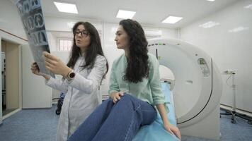 Woman doctor radiologist explains the results of CT scanning for young female patient, showing the snapshot with images, observing and analyzing CT scan in modern clinic beside modern CT scanner 19 video