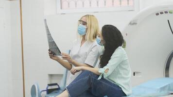 Woman doctor radiologist explains the results of CT scanning for young female patient, showing the snapshot with images, observing and analyzing CT scan in modern clinic beside modern CT scanner 15 video
