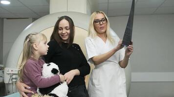 Woman doctor radiologist explains results of MRI scanning for woman with her child, showing the snapshot with images, observing and analyzing mri scan in modern clinic beside modern closed-type MRI video