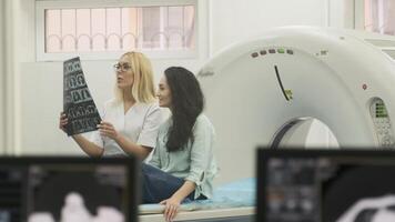 Woman doctor radiologist explains the results of CT scanning for young female patient, showing the snapshot with images, observing and analyzing CT scan in modern clinic beside modern CT scanner video