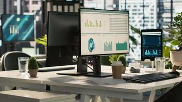 Modern office workspace desk with computer monitors displaying graphs, charts and data analytics, surrounded by plants and paperwork with city environment background, handheld camera shot video